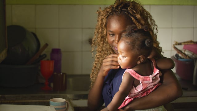 Person living with rheumatic heart disease holds her baby in her house's kitchen in Mozambique.