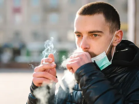 Closeup man with mask during COVID-19 pandemic coughing and smoking a cigarette at the street