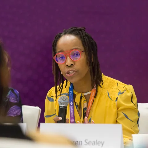 Caribbean woman in yellow dress talking into microphone at Global NCD Forum